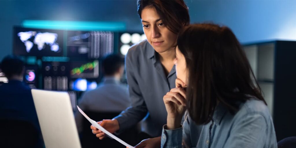 Two female colleagues (one using a computer) reference their cybersecurity documentation.