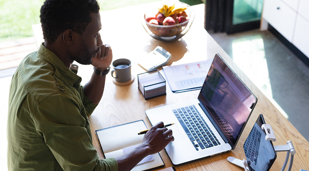 Happy african american man standing at table in kitchen, using laptop and tablet